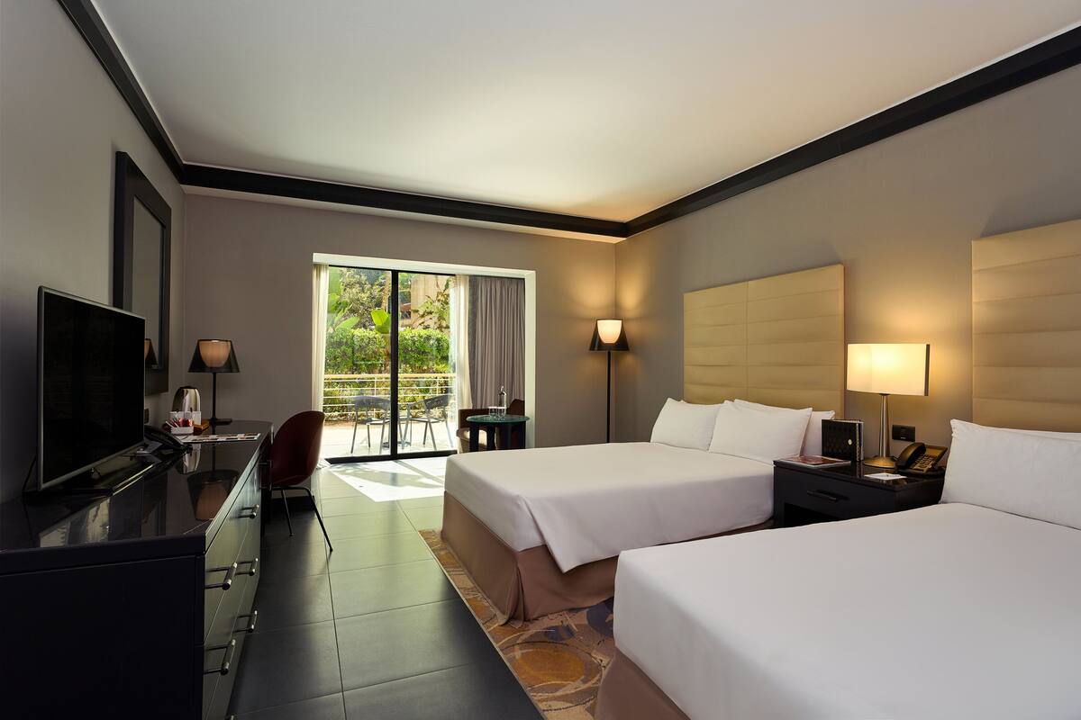 A hotel room with two beds, a desk with a TV, a chair, a small table, and a view of the outdoors.