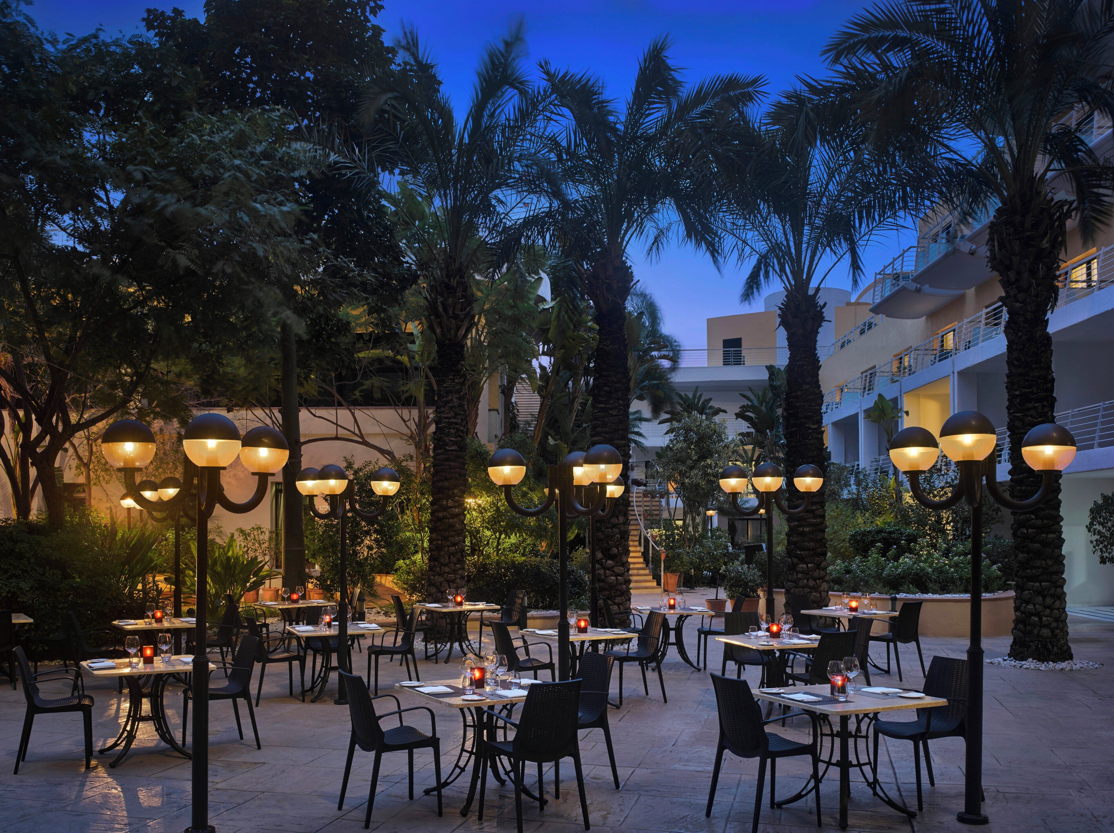 An outdoor dining area with tables and chairs, surrounded by palm trees and a building in the background.