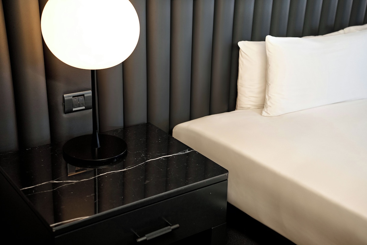 A neatly made bed with white linens, a black bedside table with a lamp, and a power outlet.