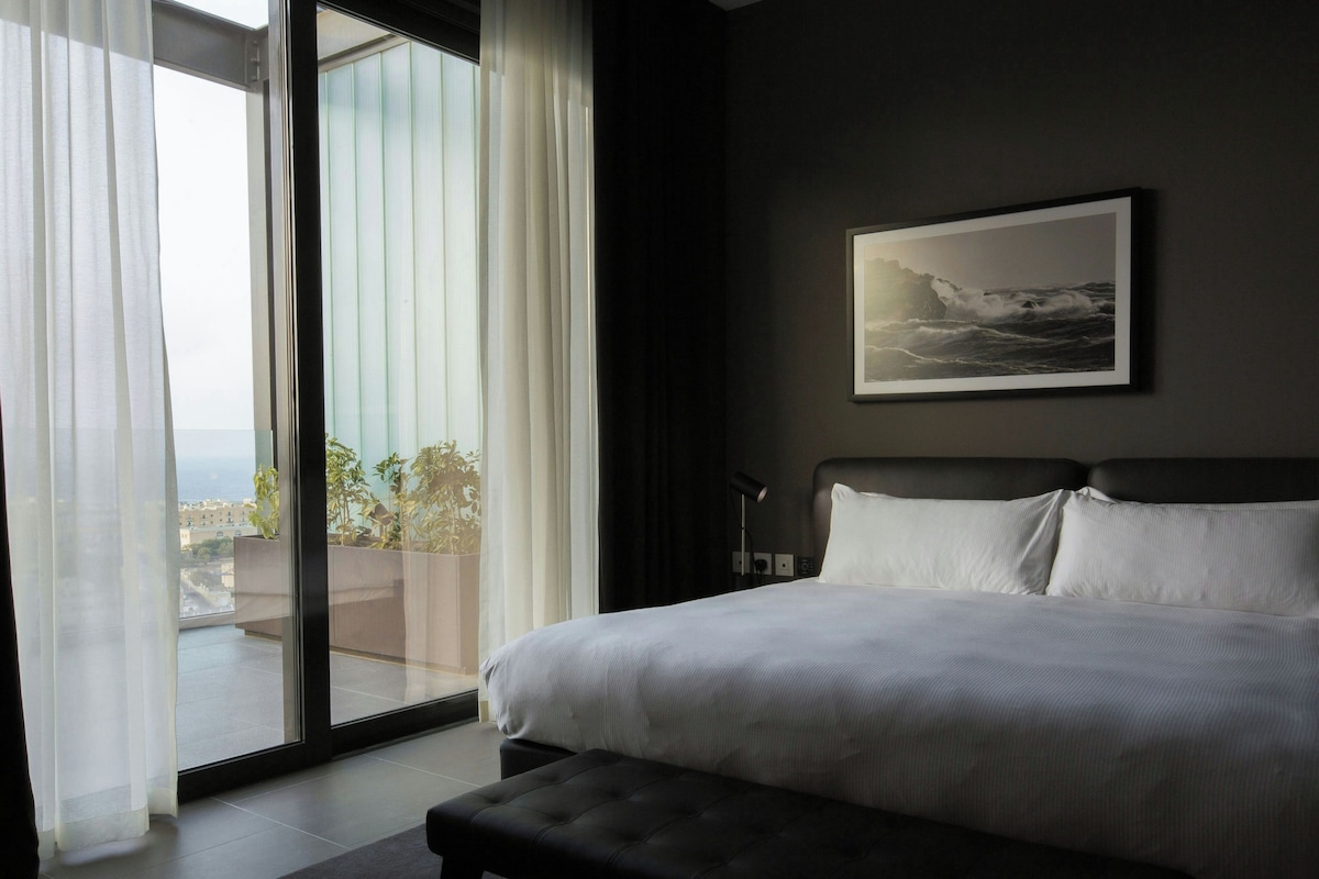 Habitación de hotel con una cama grande, vista de la ciudad a través de un ventanal y un cuadro en la pared.