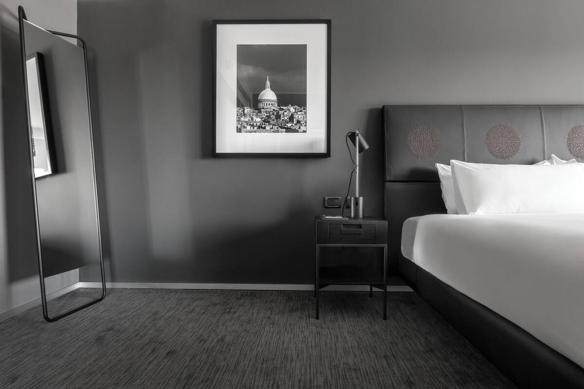 Quarto de hotel moderno com uma cama grande, uma fotografia emoldurada da paisagem urbana na parede, uma mesa de cabeceira com abajur e cabeceira de cor escura.