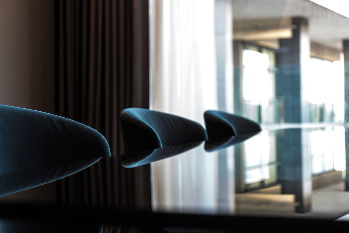 A row of dark-colored chairs with a modern design, positioned in a well-lit indoor space with a large window and a glass barrier.