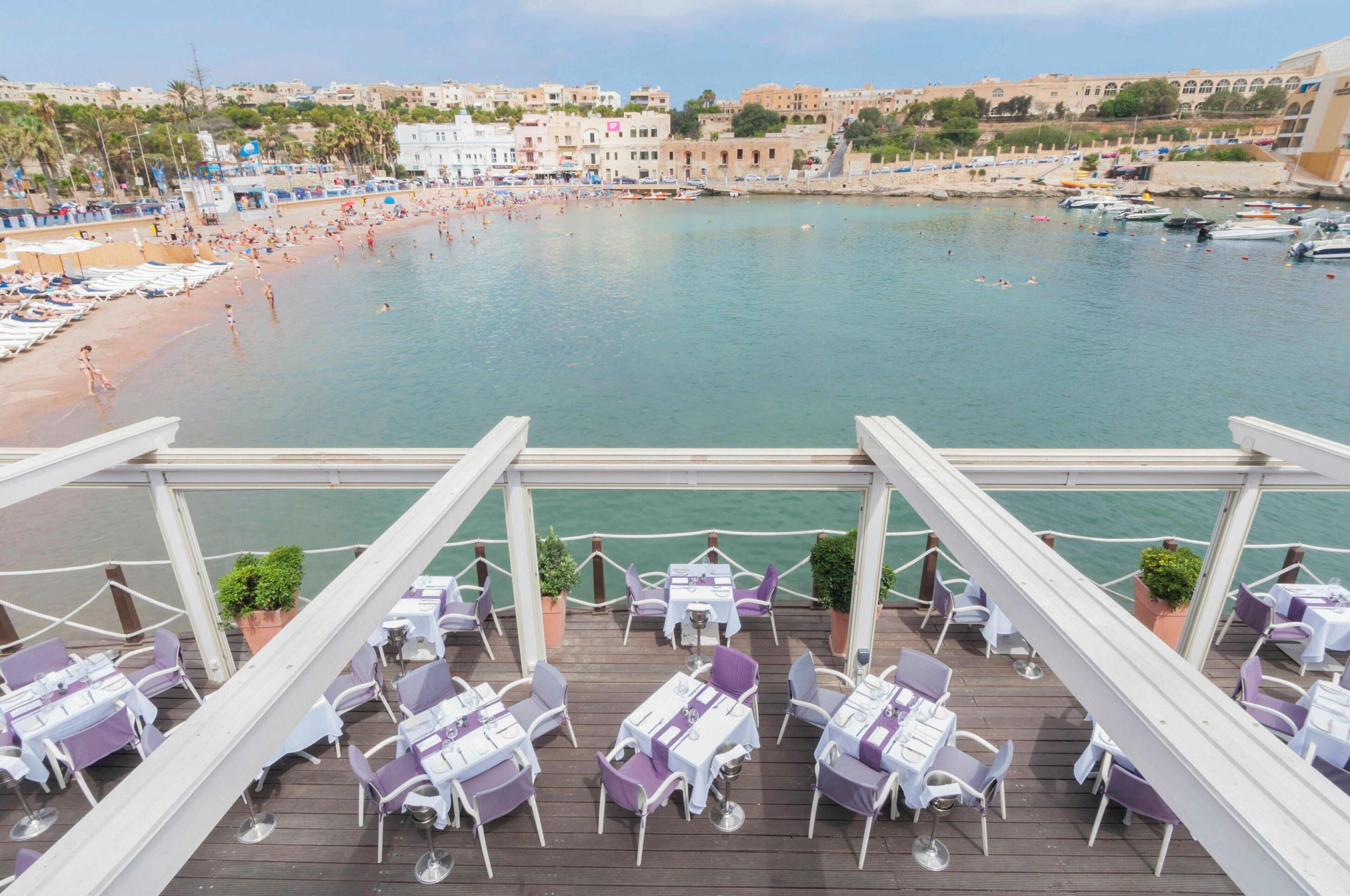 Terraza frente al mar con mesas y sillas, con vista a una playa y un puerto deportivo.