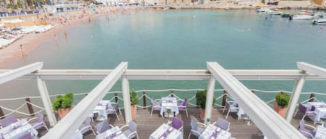 A seaside terrace with tables and chairs, overlooking a beach and marina.