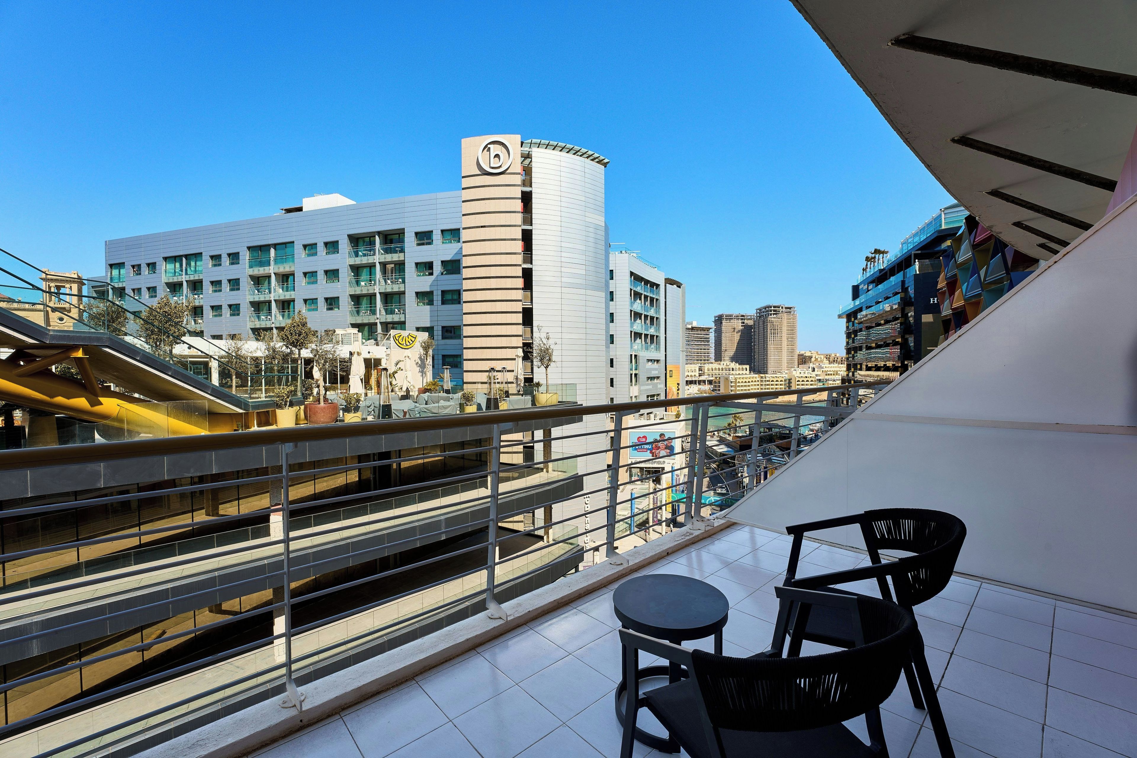Ein Balkon mit Blick auf eine Stadtlandschaft, darunter ein modernes Gebäude mit einer runden Spitze und ein Hotel mit einer markanten blau-weißen Fassade.