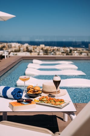 A poolside dining area with a table set for a meal, a drink, and a book. - InterContinental Malta by IHG (St. Julian's)
