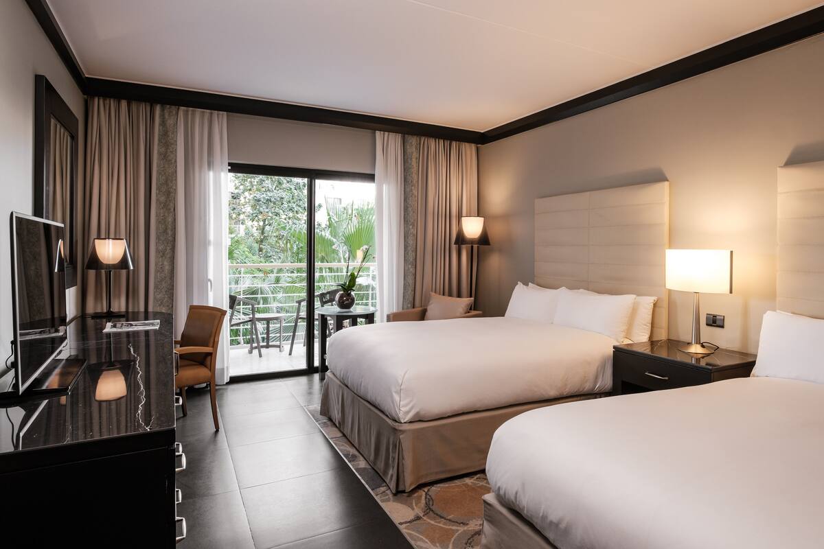 Quarto de hotel com duas camas, uma escrivaninha, uma cadeira e varanda com vista para o verde.