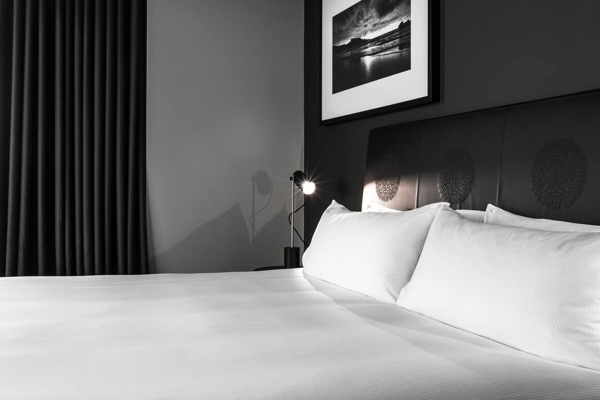 A neatly made bed with white linens, a headboard with decorative panels, a framed picture above the bed, and a floor lamp to the left.