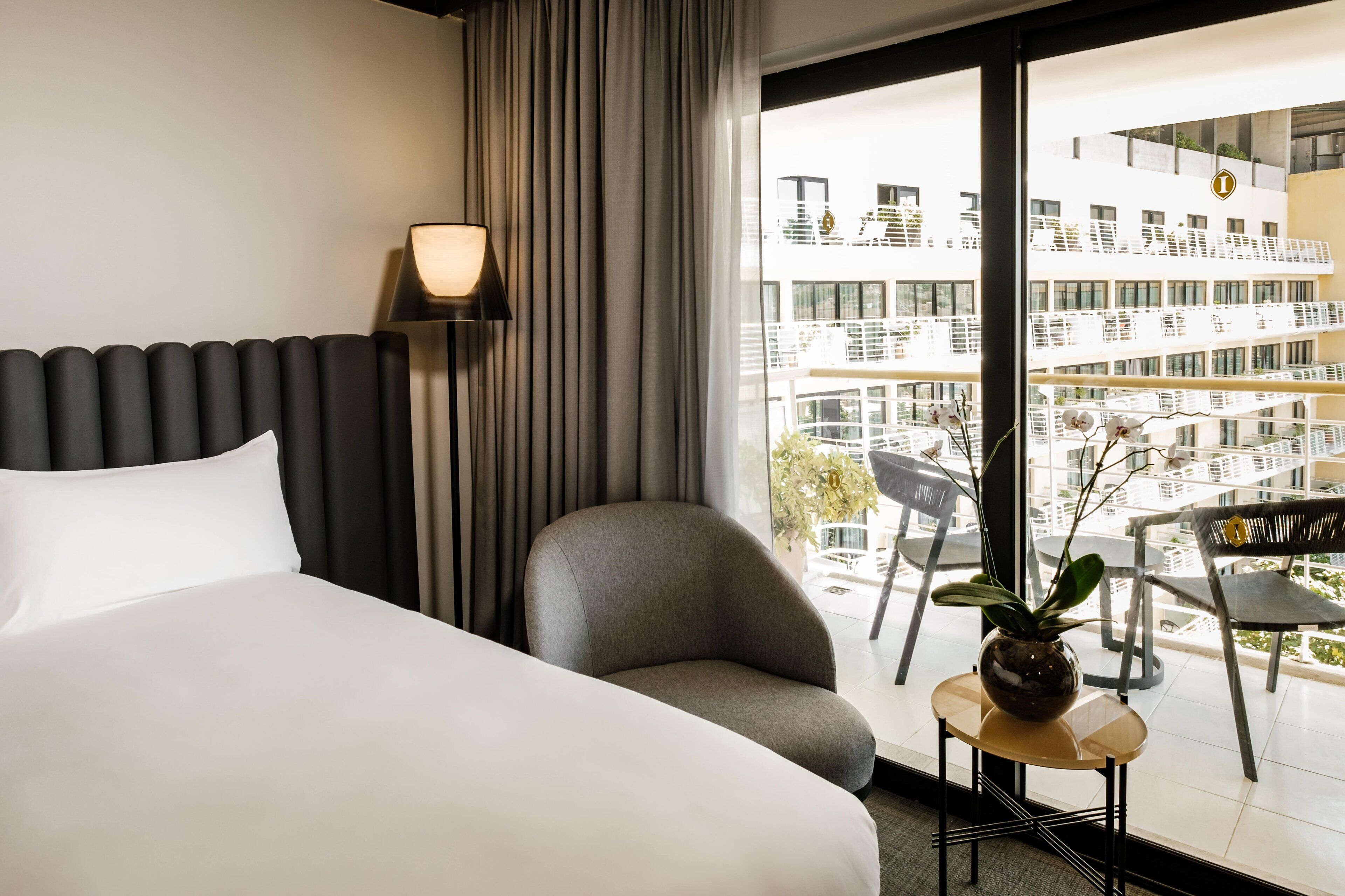 Ein Hotelzimmer mit Bett, Sessel, kleinem Tisch mit Pflanze, Lampe und Blick auf ein modernes Gebäude durch große Fenster.