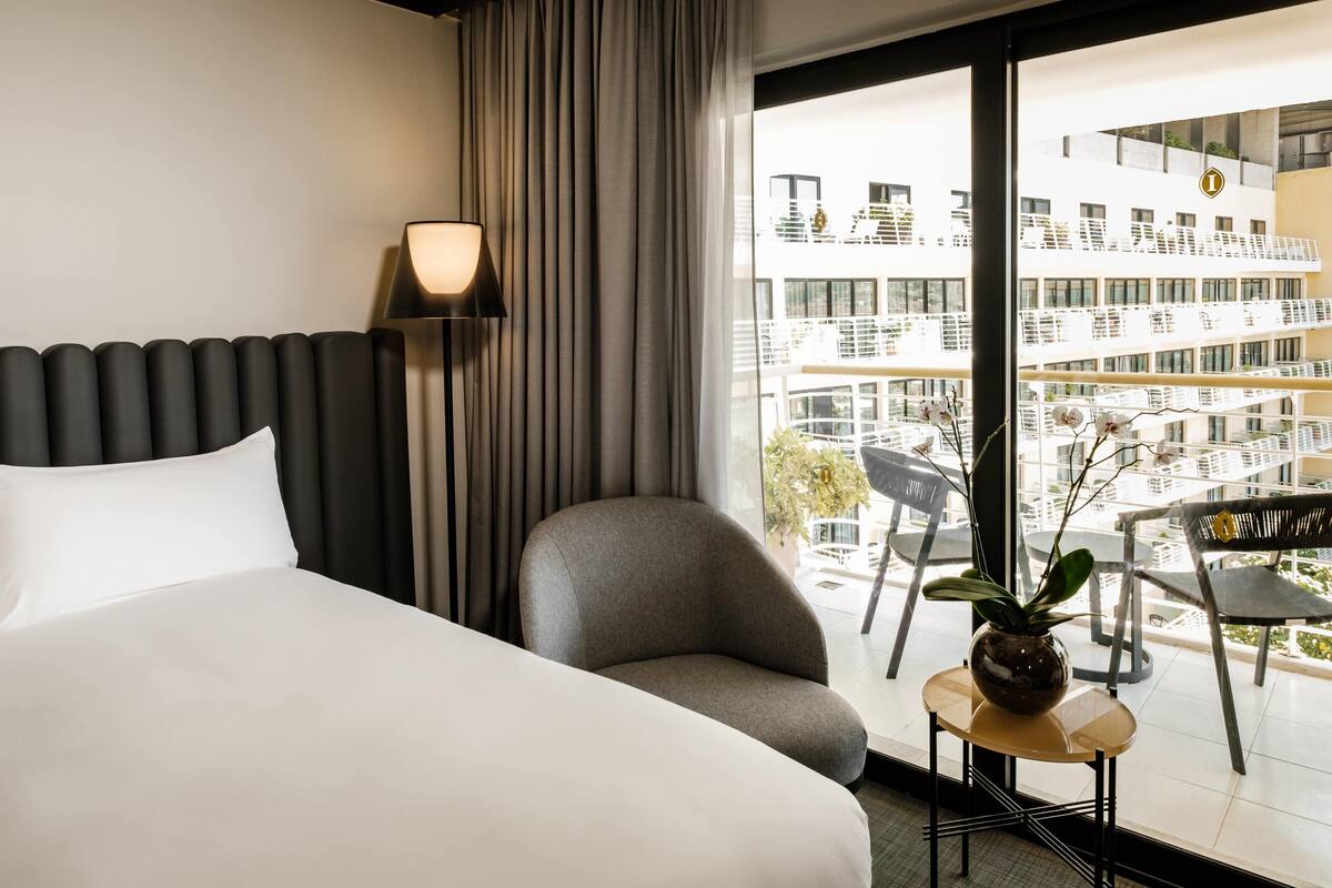 Quarto de hotel com uma cama, uma cadeira, uma mesa pequena com uma planta, um abajur e vista para um edifício moderno através de grandes janelas.