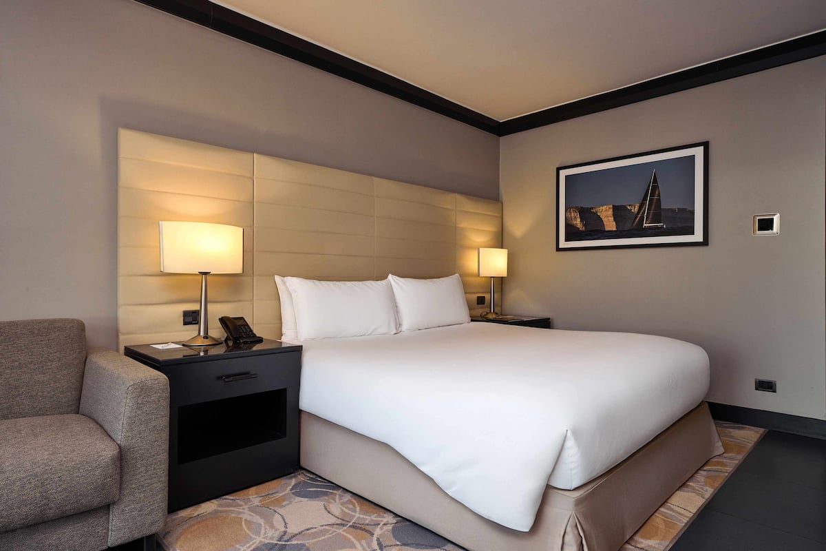 Una camera d'albergo con un letto grande, due comodini con lampade, una sedia e un quadro appeso al muro.