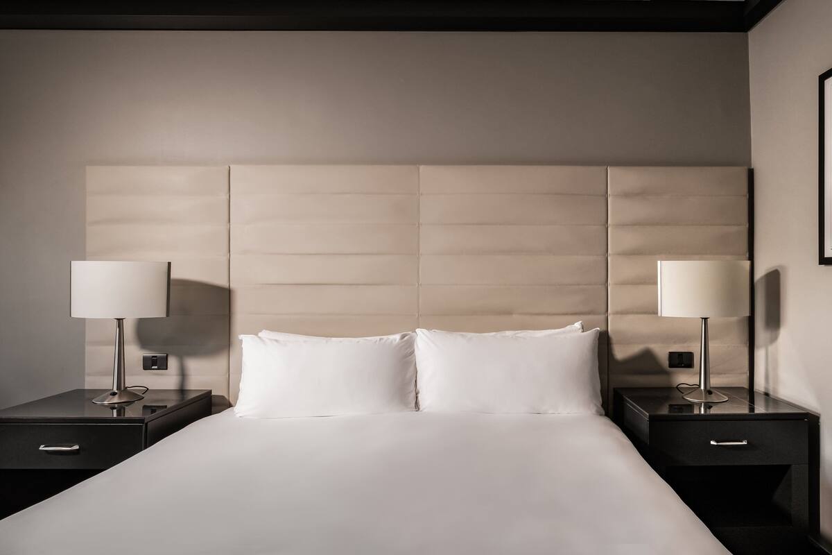 Cama bem arrumada com lençóis brancos, com duas mesas de cabeceira com luminárias, em quarto de hotel moderno com cabeceira acolchoada.