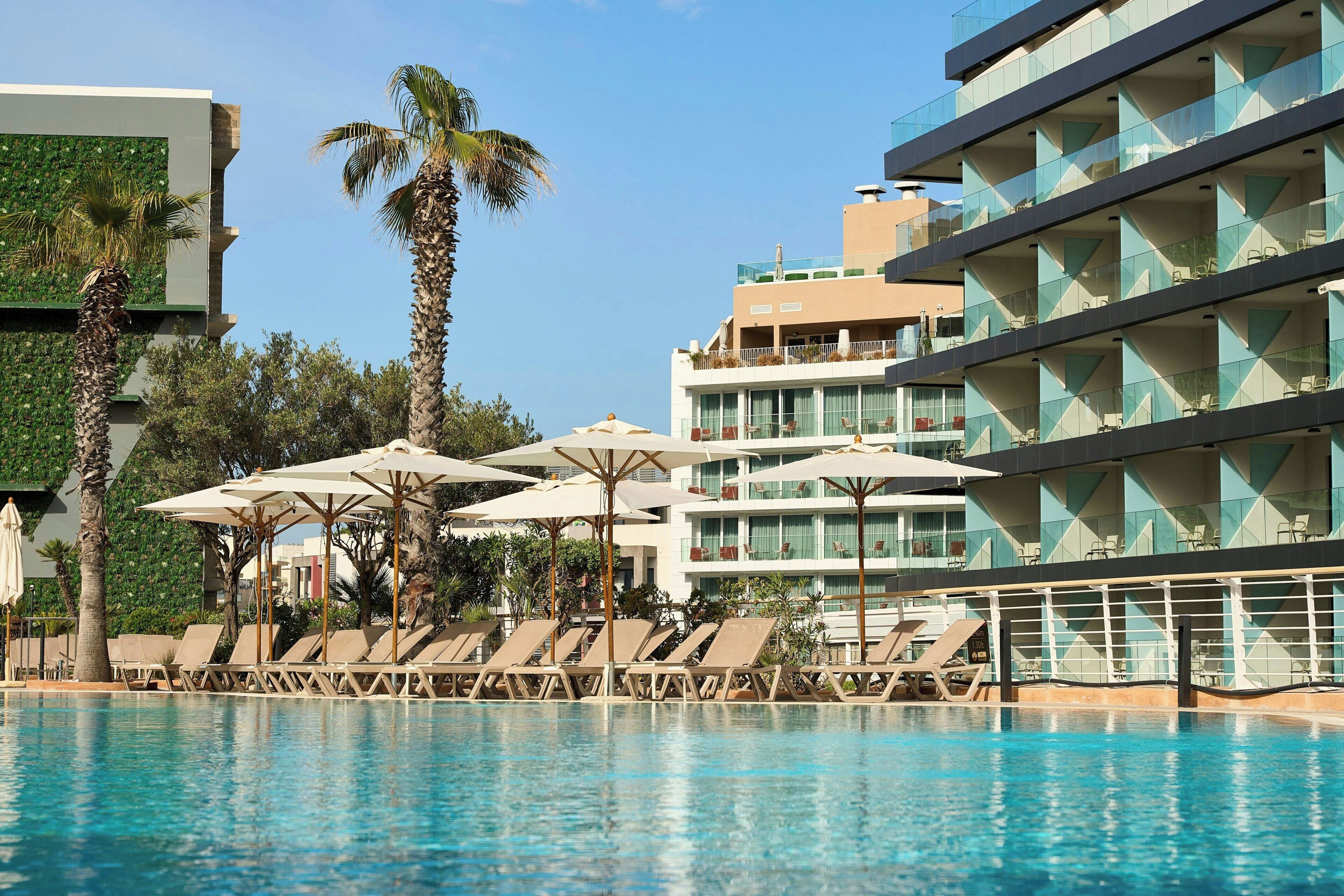 Zona de piscina de hotel con tumbonas y sombrillas, rodeada de palmeras y edificios modernos.