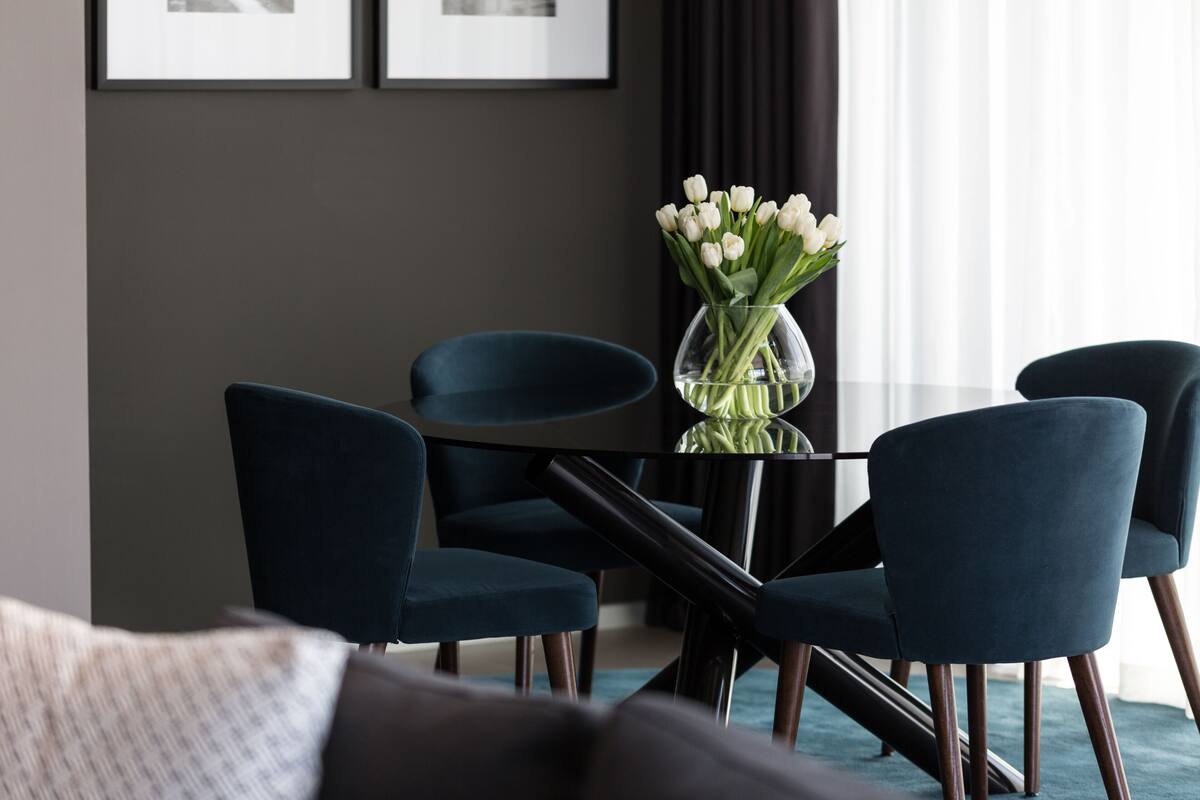 A modern hotel room with a dark blue sofa, a glass coffee table with a vase of white flowers, and a dark blue chair.