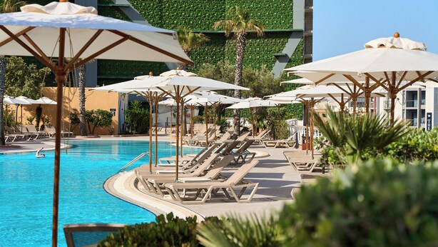 Zona de piscina con sillones de descanso y sombrillas, rodeada de vegetación y un edificio moderno con un muro verde.