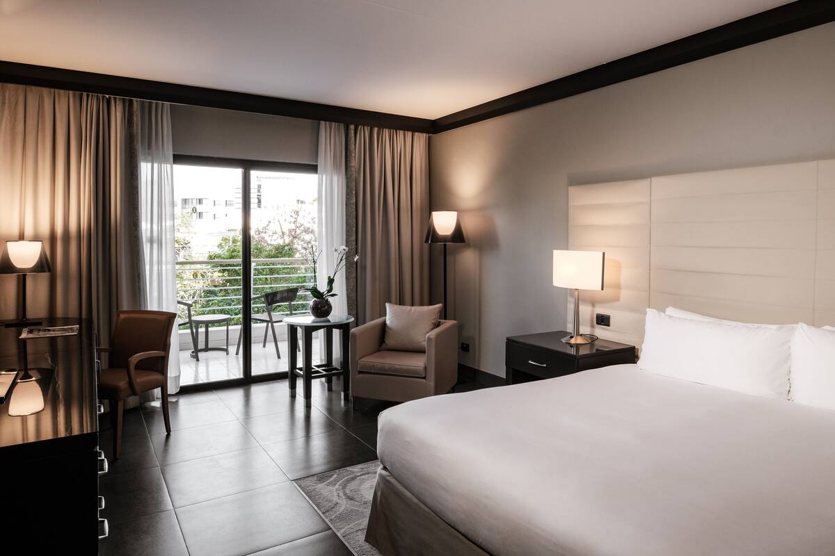 Quarto de hotel com uma cama grande, uma escrivaninha com luminária, uma cadeira e vista para uma varanda com mobiliário para áreas externas.