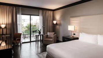 A hotel room with a large bed, a desk with a lamp, a chair, and a view of a balcony with outdoor seating.