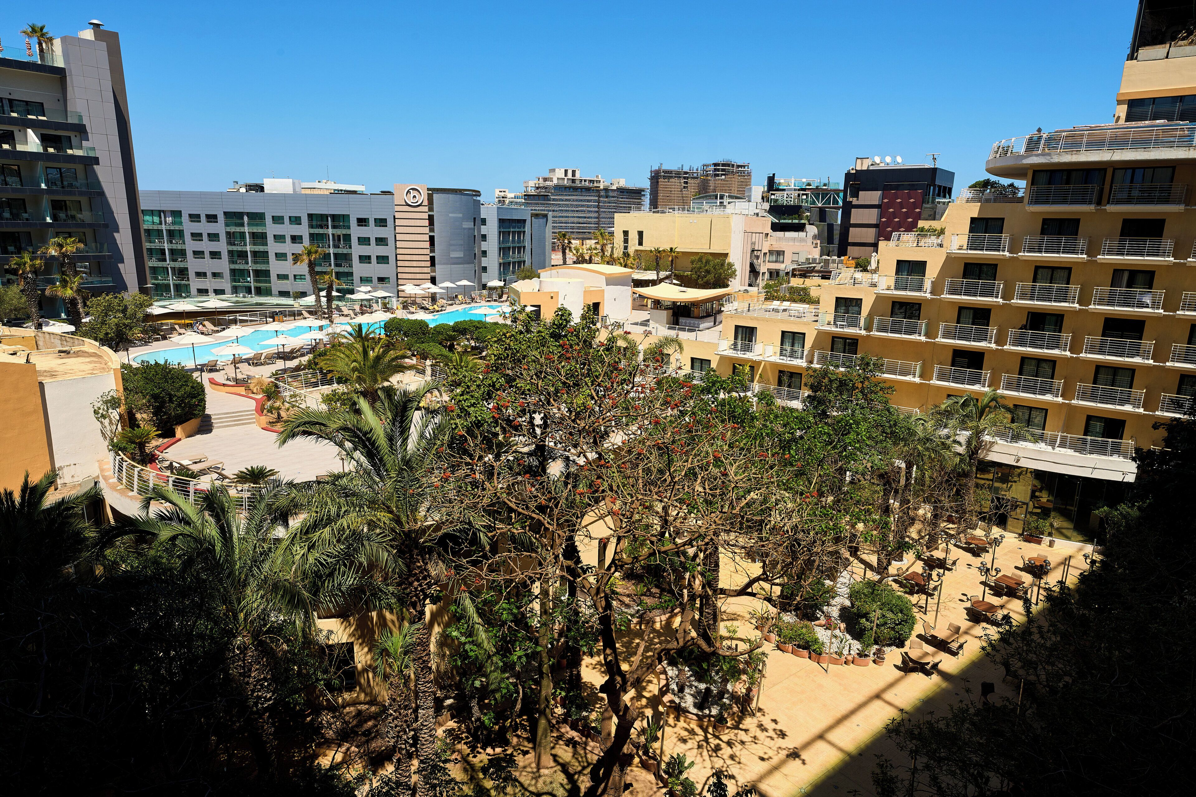 Un hotel con piscina, varios pisos y abundante vegetación alrededor.