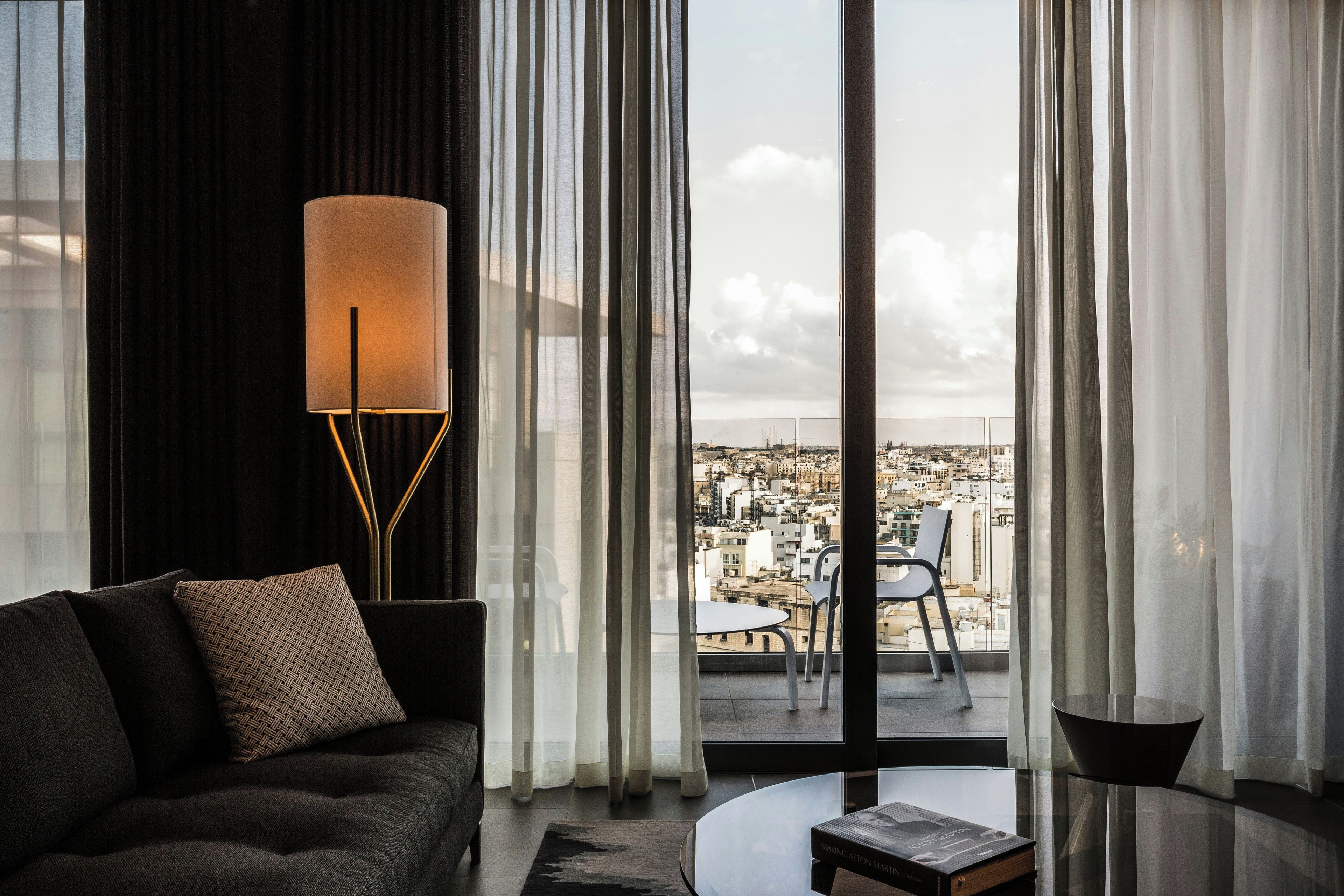 Una sala de estar moderna con un sofá, una lámpara de pie y vistas de un paisaje urbano a través de amplios ventanales.