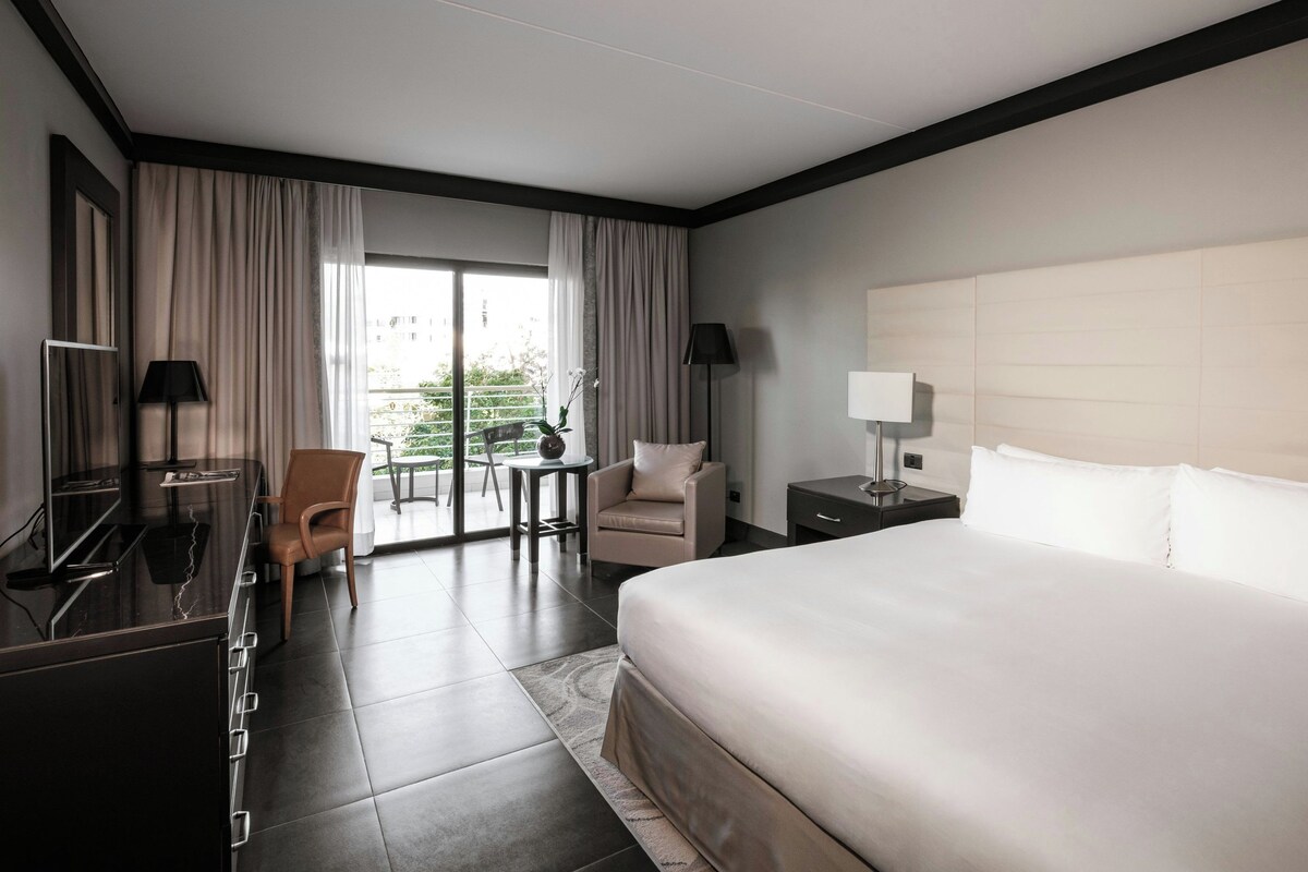 A hotel room with a large bed, a desk, a chair, a small table, and a balcony with curtains.