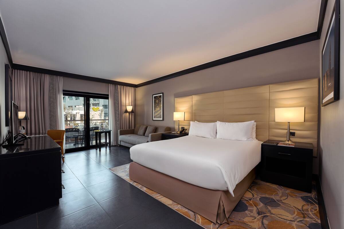 Quarto de hotel com uma cama grande, uma escrivaninha, uma cadeira, um sofá e vista da cidade pela janela.