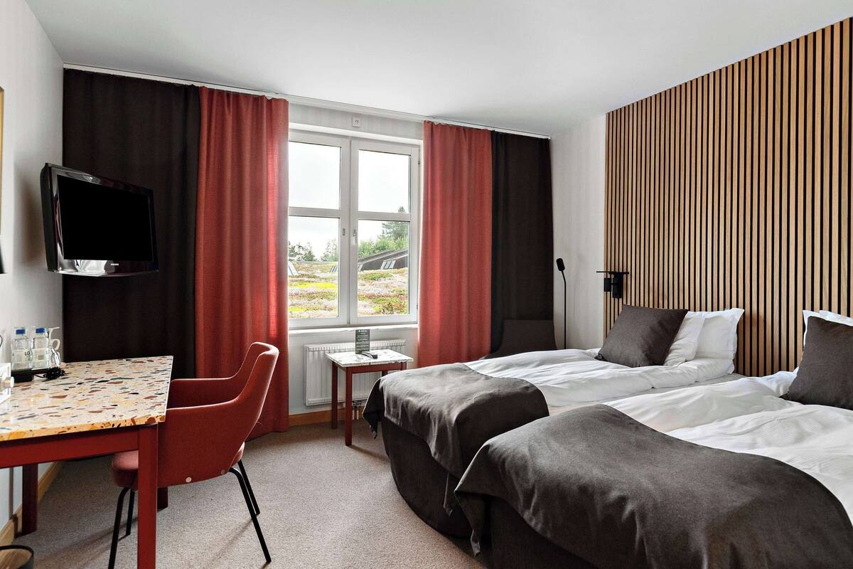 Ett hotellrum med två sängar, en TV, ett litet bord med stolar och ett fönster med gardiner.