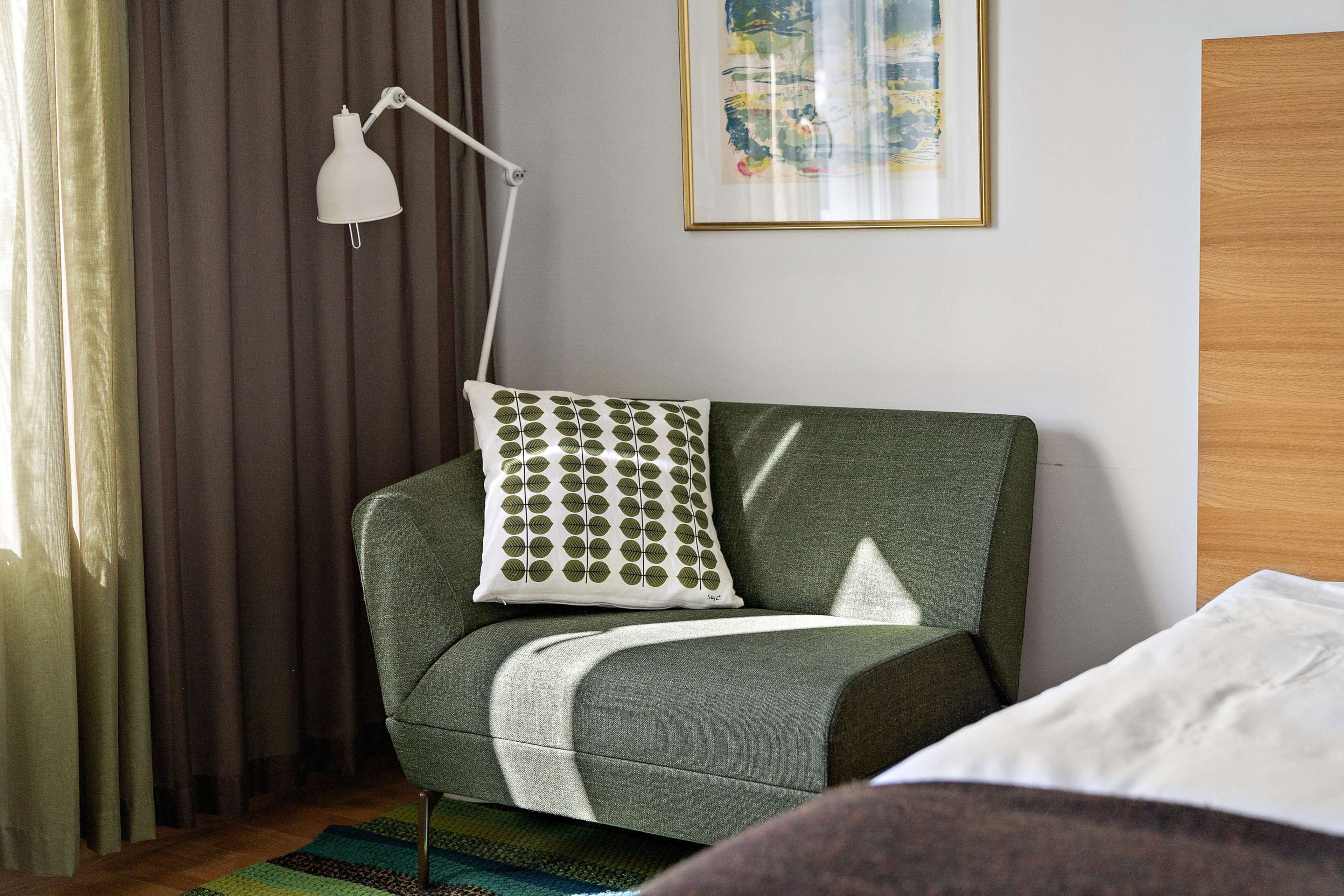 Ein Zimmer mit einem grünen Sofa, einer weißen Lampe, einem gemusterten Kissen, einem gerahmten Bild an der Wand und einer Holztür.