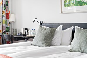 Una cama bien tendida con almohadillas estampadas, una mesita de noche con lámpara, un cuadro enmarcado en la pared y una cortina colorida.