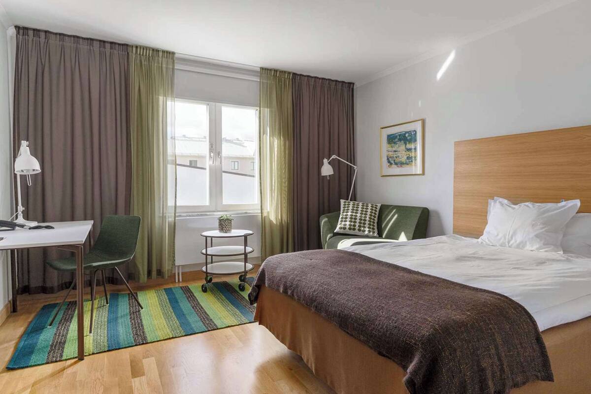 Ett hotellrum med en säng, ett skrivbord, en stol, ett litet bord och ett fönster med gardiner.