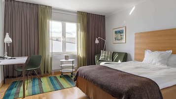 Una habitación de hotel con cama, escritorio, silla, mesita y ventana con cortinas.
