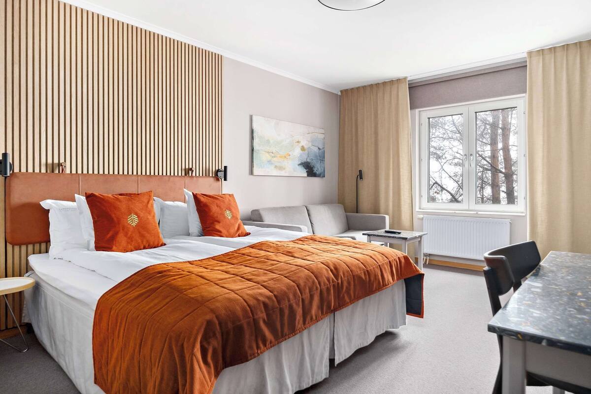 Ett hotellrum med en säng, ett skrivbord, en stol, ett fönster med gardiner och ett konstverk som hänger på väggen.
