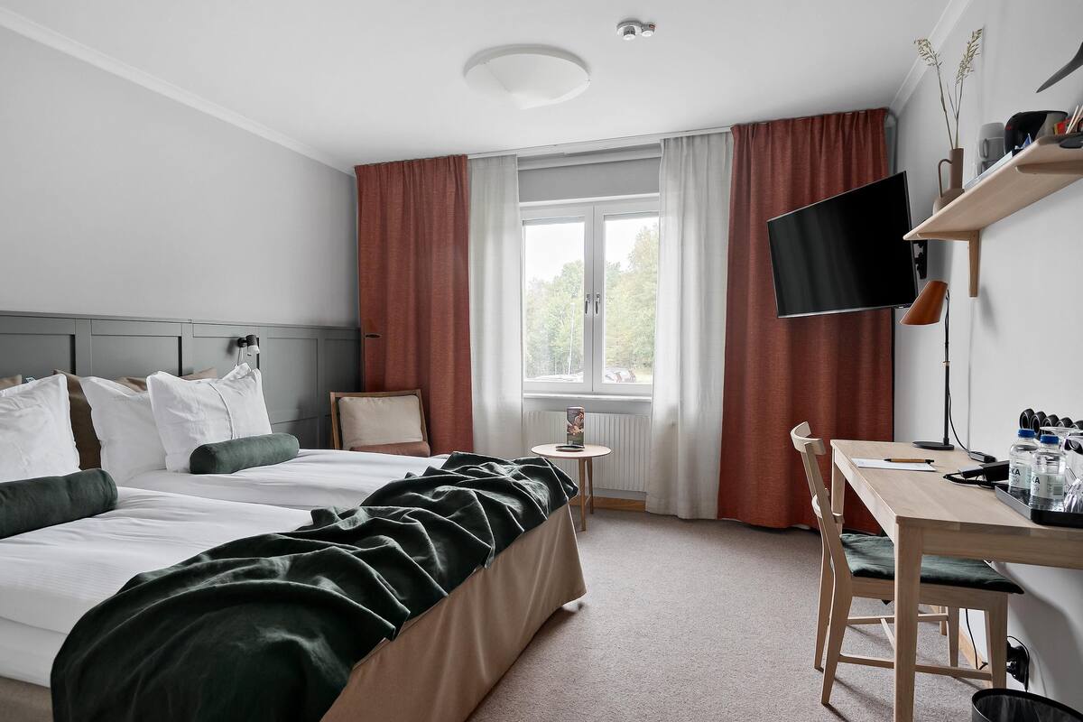 Ett hotellrum med en säng, ett skrivbord med en stol, en tv, ett fönster med gardiner och ett element.