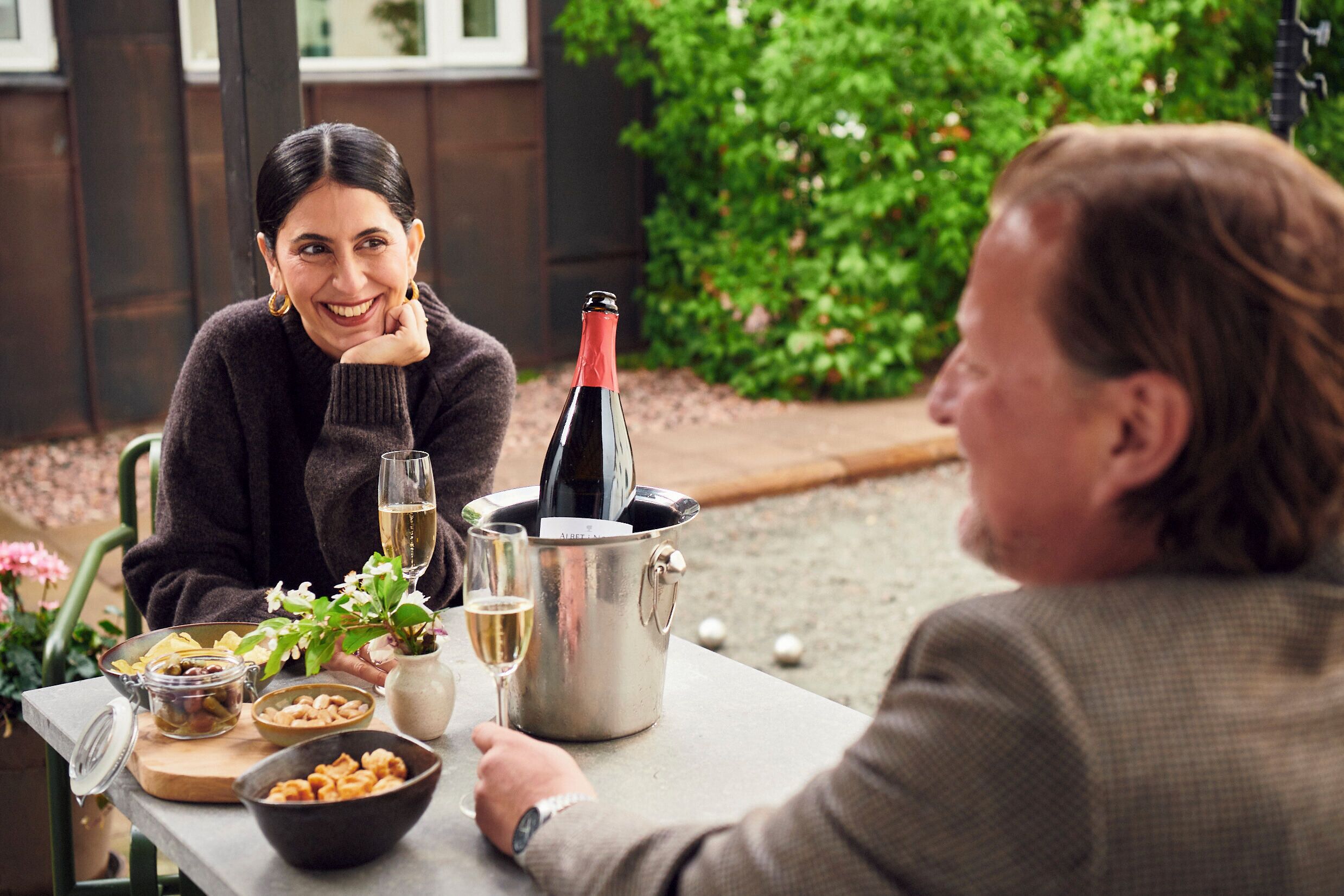Two people seated at an outdoor table with a bottle of wine, glasses, and bowls of food.