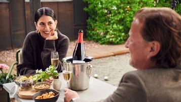 Two people seated at an outdoor table with a bottle of wine, glasses, and bowls of food.