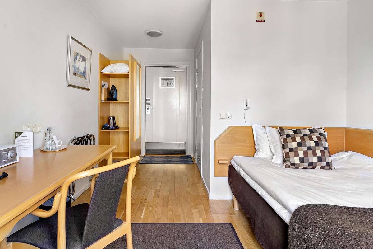 Ett hotellrum med en säng, ett skrivbord, en stol, en kaffebryggare och en inramad tavla på väggen.