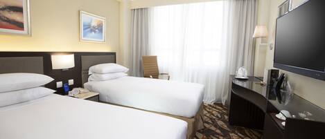 Een hotelkamer met twee bedden, een bureau, een flatscreen televisie, een stoel en een groot raam met gordijnen.