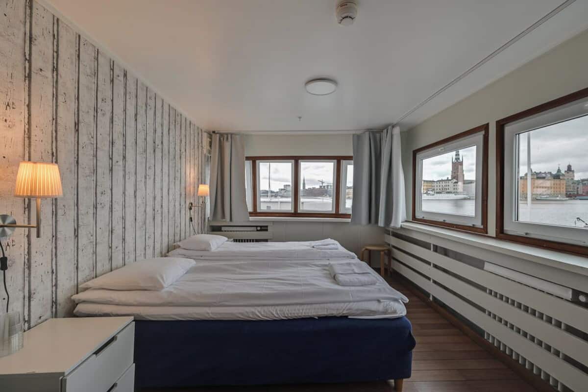 A hotel room with a large bed, a desk with a lamp, and a view of a cityscape through large windows.
