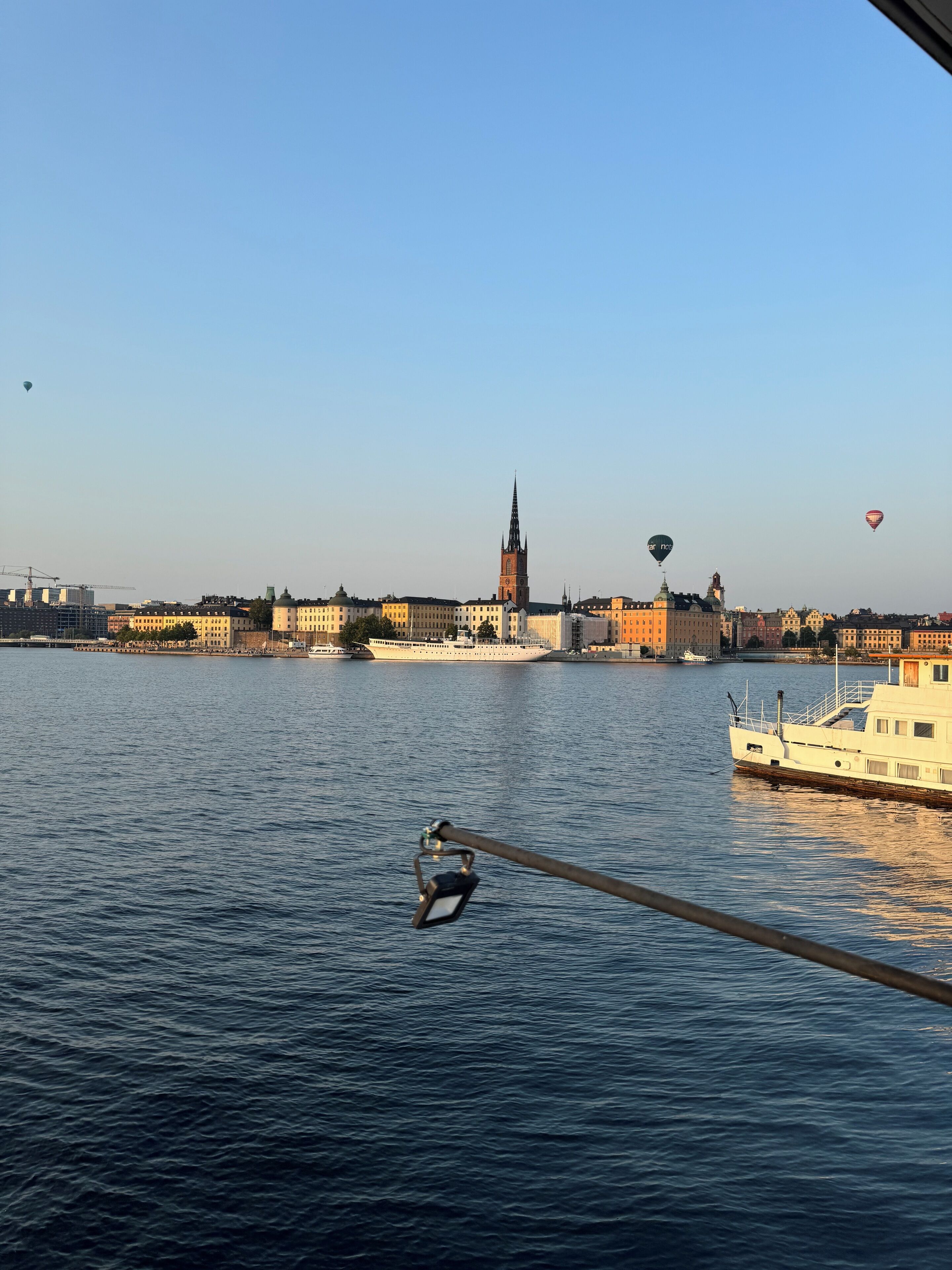 A river with a boat docked on the right, a city skyline in the background, and hot air balloons in the sky.