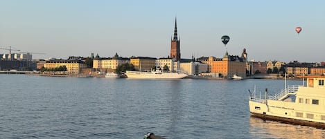 A river with a boat docked on the right, a city skyline in the background, and hot air balloons in the sky.