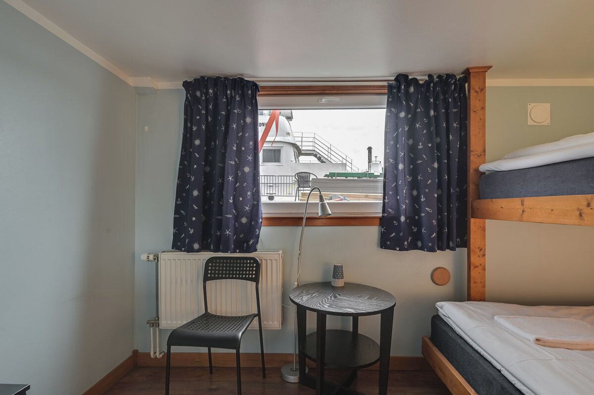 A small room with a bunk bed, a desk with a chair, a small table, and a window with curtains.