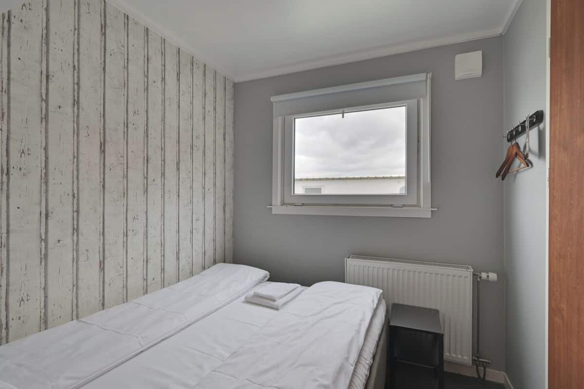 A small, single-bed room with a white bed, a window, a radiator, and a wooden wall.