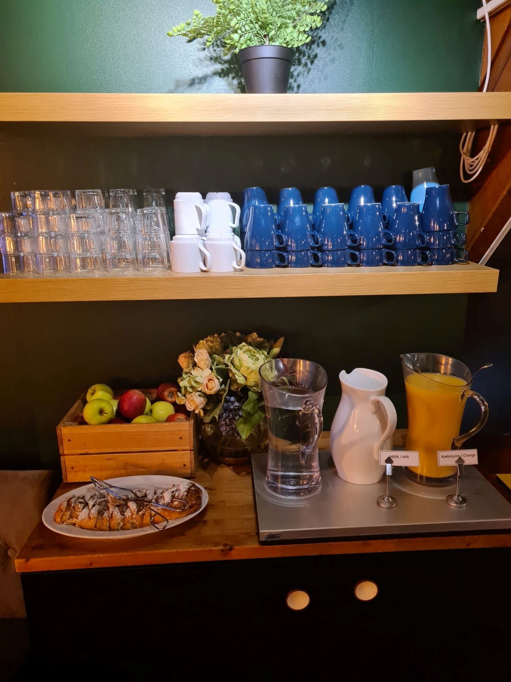 A breakfast bar with stacked cups, a pitcher of water, a jug of orange juice, a plate of pastries, and a wooden crate of apples and flowers.