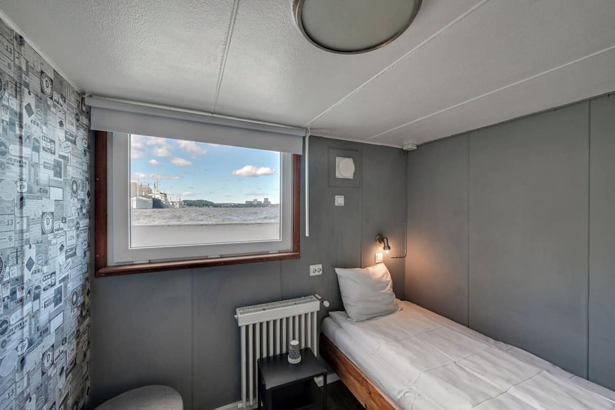 A small, compact room with a single bed, a radiator, a small table, and a window with a view of a distant cityscape.