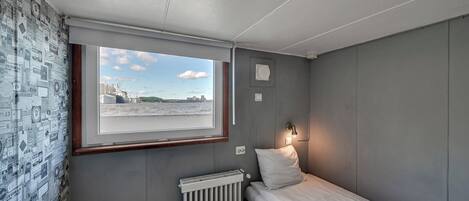 Una habitación pequeña y acogedora con una cama individual, un radiador, una mesita y una ventana que ofrece vistas a un paisaje urbano lejano.