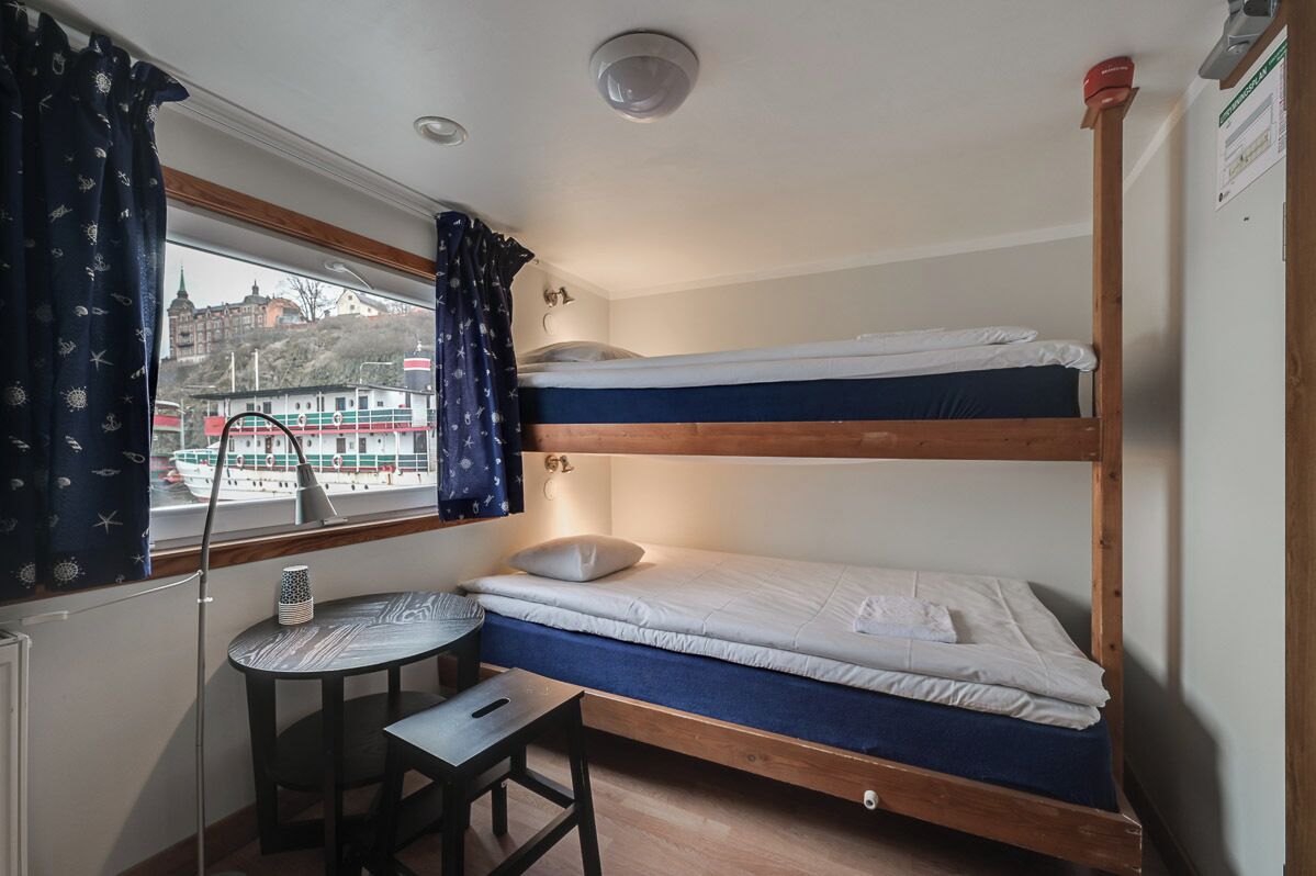 A bunk bed room with a table and chair, a window with curtains, and a view of a train and buildings outside.