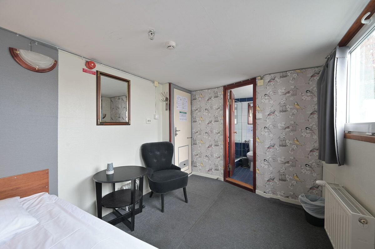 A hotel room with a bed, a small table with a cup, a chair, a mirror, and a door leading to another room.