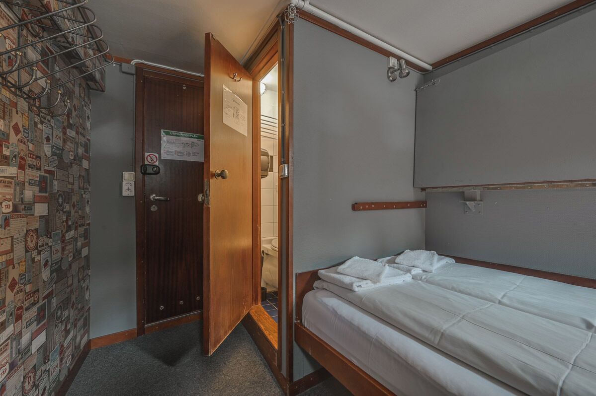 A small, single-bed room with a wooden door, a towel on the bed, and a wall with a bulletin board.