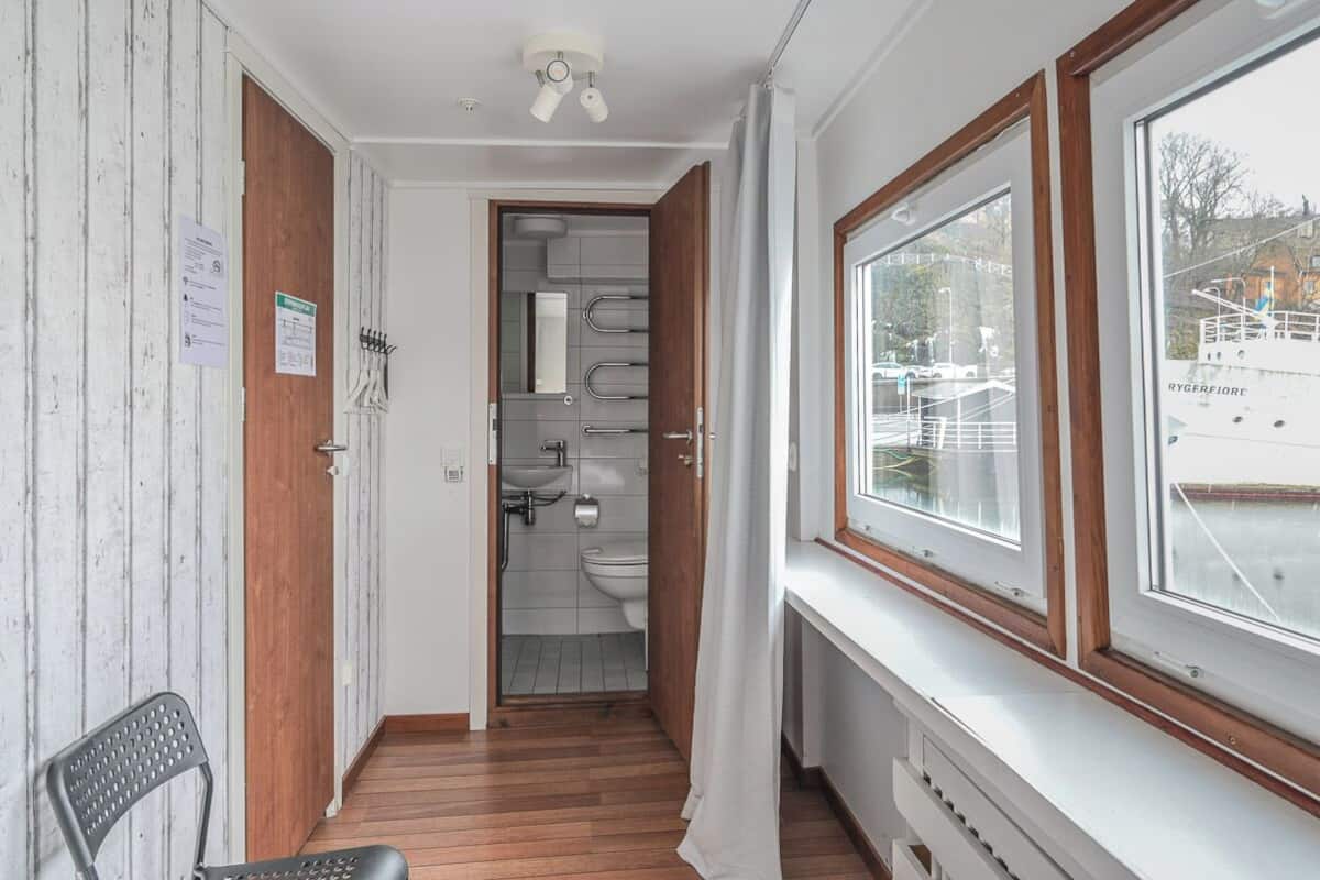 A narrow hallway leading to a bathroom with a toilet, sink, and shower. A chair is placed against the wall, and a window provides a view of a boat outside.