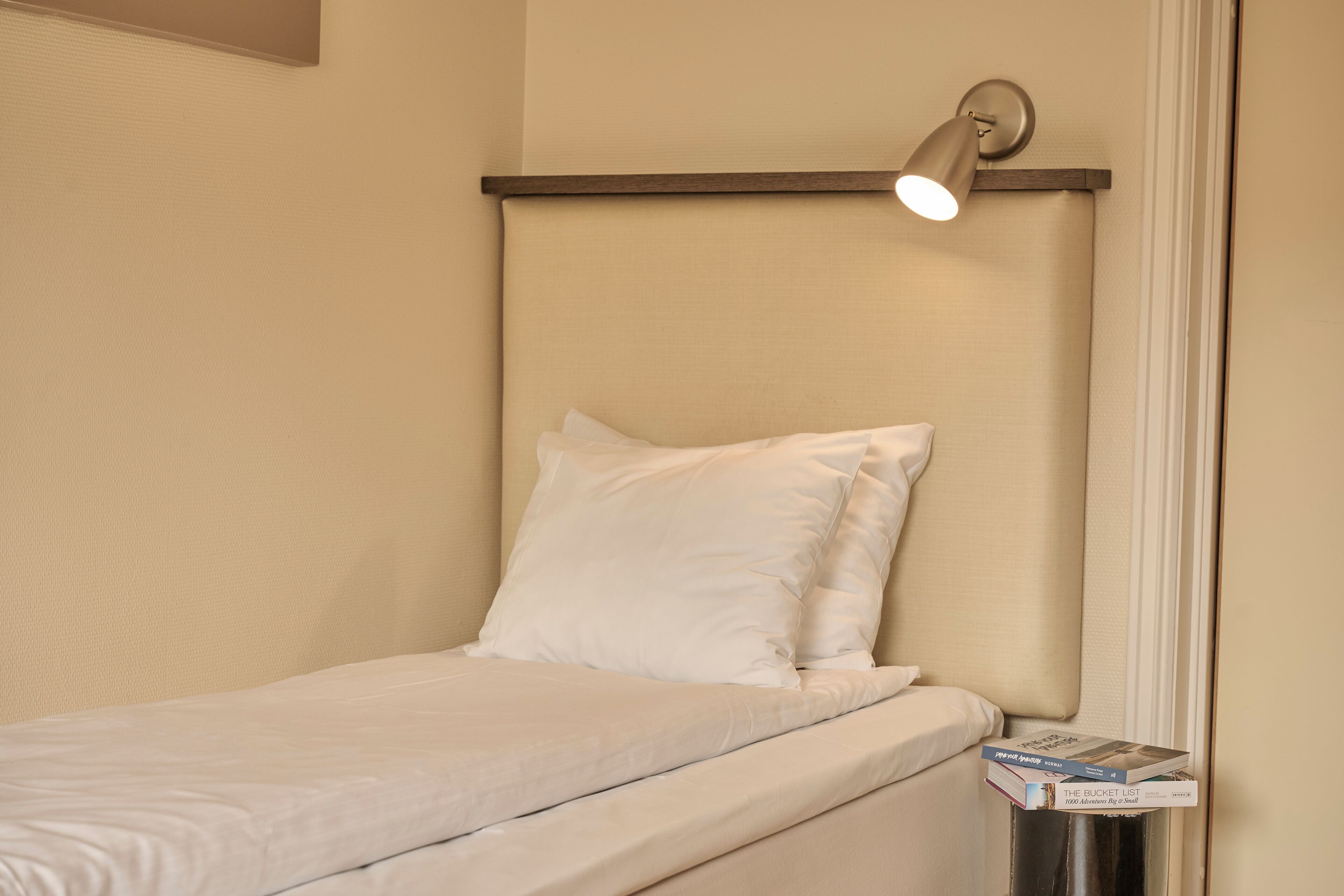 Un lit bien fait, avec du linge de lit blanc et des oreillers, une lampe de chevet et une pile de livres sur une petite table placée à côté du lit.