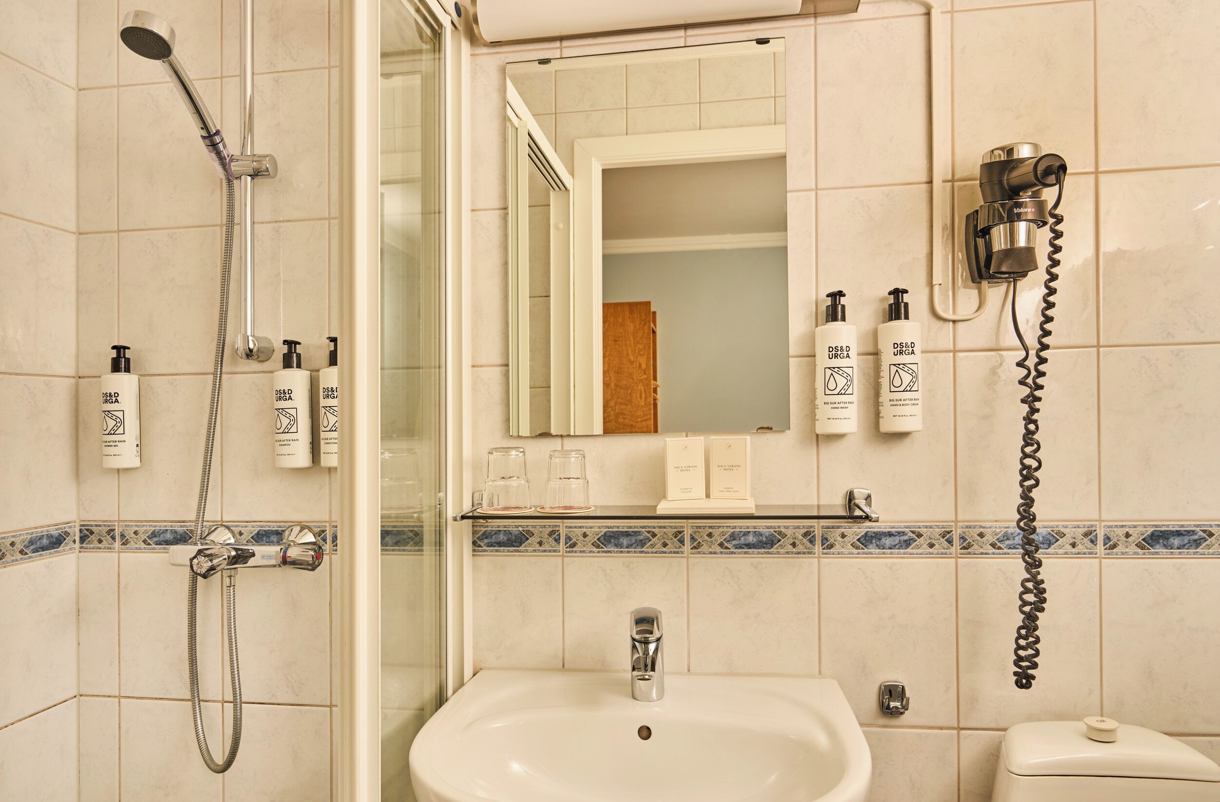 Une salle de bain avec une douche, un lavabo et des toilettes. On y trouve des articles de toilette accrochés au mur et un miroir au-dessus du lavabo.